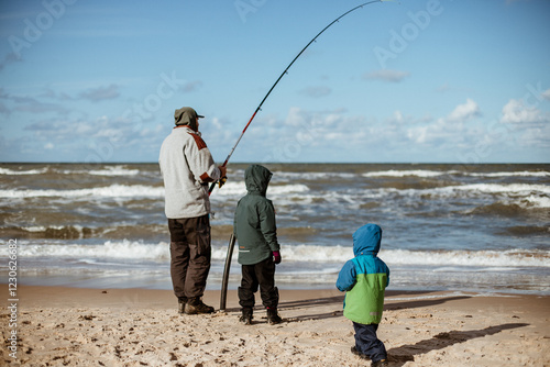 A father with two young sons fishing for flounders at the seaside on a sunny day, with waves in the sea. The view is from behind. Grain effect.