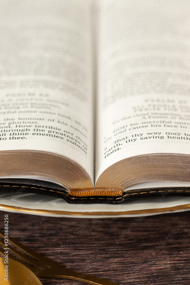 Open holy bible book on plate with golden fork and spoon on wooden table. Close-up. Spiritual food and Christian fasting concept. Open holy bible book on plate with golden fork and spoon on wooden table. Close-up. Spiritual food and Christian fasting concept.