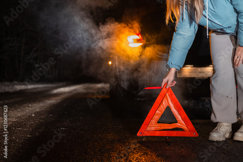 On a dark winter road, a woman places an emergency triangle behind her broken-down car. The scene captures the cold atmosphere with visible exhaust and illuminated taillights.