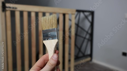 A hand holds a 2 inch natural bristle paintbrush. A wooden gate with vertical slats and a black geometric metal frame is visible in the indoor setting.
