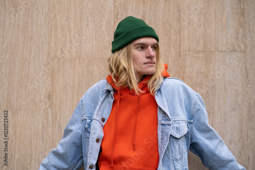 Blonde long-haired hipster guy in colorful trendy stylish outfit, posing, leaning against wall in city. Carefree fashionable male in orange hoodie and jean, representative of generation z photo