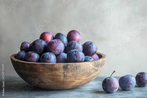 A wooden bowl filled with ripe plums, some additional plums sit beside the bowl on a grey surface.