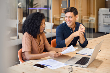 Two young diverse business people using laptop and talking at office meeting. Busy happy team professional employees working together on computer discussing online project at work. Candid photo.