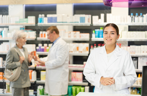 Positive young girl pharmacist posing in chemist's shop with large assortment of goods