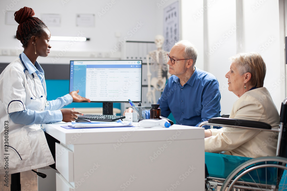 Obraz premium Elderly female wheelchair user and her husband speaking with african american doctor during medical consultation. Physician offering reassurance about diagnosis to caucasian senior couple.