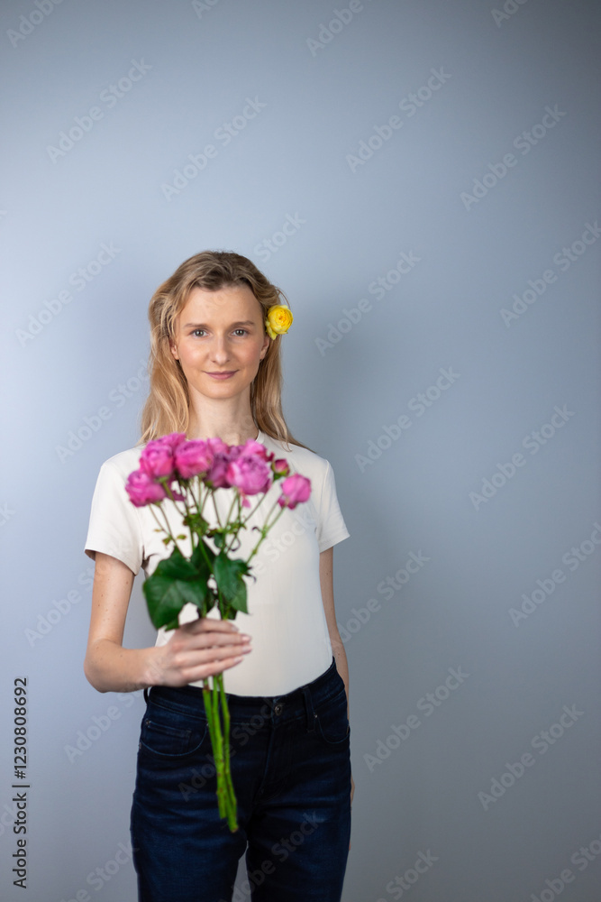 on a light gray background a young blonde girl in a light T-shirt in her hands pink peony roses March 8 smile on her face bud in her hair looking at the camera on a light gray background a young blonde girl in a light T-shirt in her hands pink peony roses March 8 smile on her face bud in her hair looking at the camera