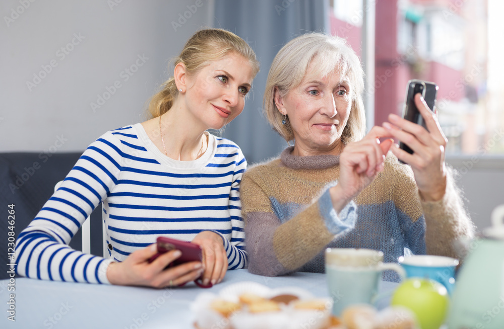 Mature woman with her adult daughter, sitting at a table in a room, looking at photos on a mobile phone Mature woman with her adult daughter, sitting at a table in a room, looking at photos on a mobile phone