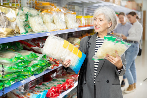 Mature female in shop carefully examines information about product on label packaging. Elderly woman customer chooses and buys pack of Chinese noodles at hypermarket photo