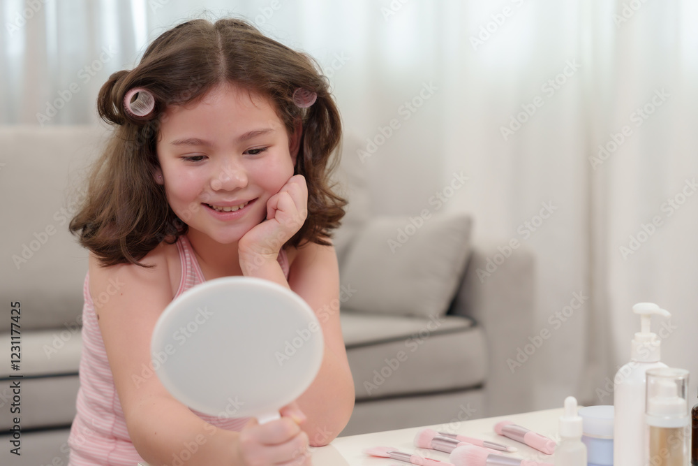 Cute Caucasian girl who loves beauty looking into a handheld mirror with a happy expression. Hair styled with curlers, sitting at a white table with makeup brushes and skincare products. Cute Caucasian girl who loves beauty looking into a handheld mirror with a happy expression. Hair styled with curlers, sitting at a white table with makeup brushes and skincare products.