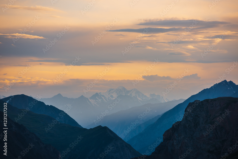 Deep gorge between layered silhouettes of sharp rocky ridge spurs under huge snowy mountain peak in golden cloudy sky. Sheer crags of mount valley under giant ice top under gold sunset color clouds. Deep gorge between layered silhouettes of sharp rocky ridge spurs under huge snowy mountain peak in golden cloudy sky. Sheer crags of mount valley under giant ice top under gold sunset color clouds.