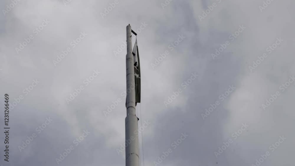 A sizeable Pakistani flag flying atop a tall pole, captured from a ground-level perspective against a backdrop of a cloudy sky. A sizeable Pakistani flag flying atop a tall pole, captured from a ground-level perspective against a backdrop of a cloudy sky.