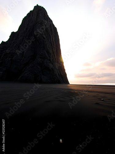 View of a high cliff rising on a beach with black volcanic sand. In the background is the setting sun, with warm rays penetrating the rock. A serene and deserted beach without people. photo