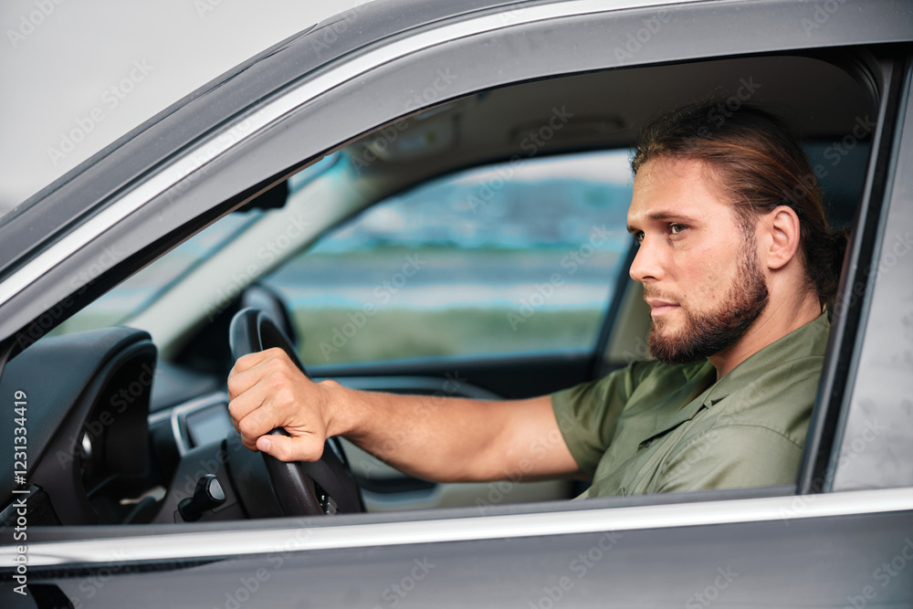 Male driver with a serious expression sitting in a car, looking out of the window Urban environment in the background Male driver with a serious expression sitting in a car, looking out of the window Urban environment in the background