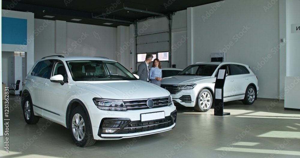 A car dealership manager in formal clothes talks to a customer and shows him a car standing in the showroom A car dealership manager in formal clothes talks to a customer and shows him a car standing in the showroom
