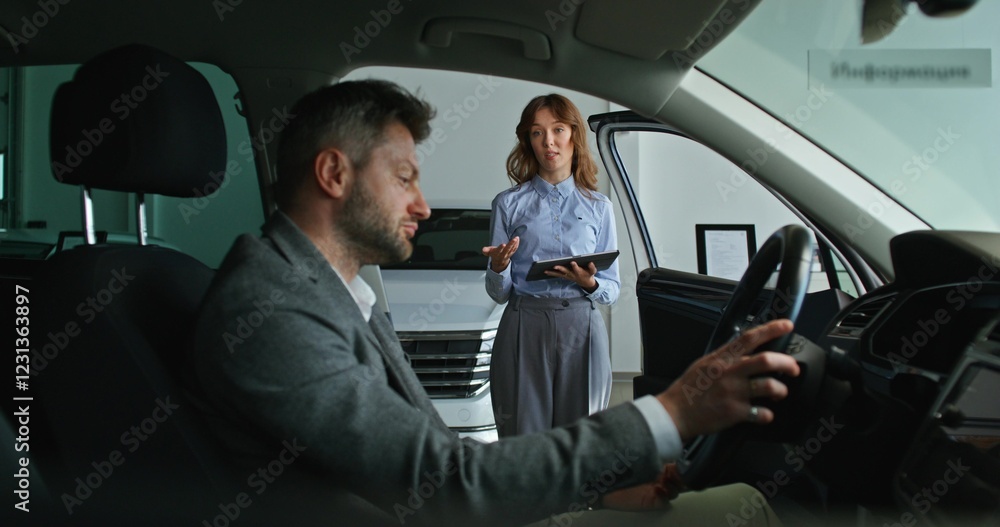 The buyer is sitting in the car and talking to the manager. A woman with a tablet in her hands stands next to him The buyer is sitting in the car and talking to the manager. A woman with a tablet in her hands stands next to him
