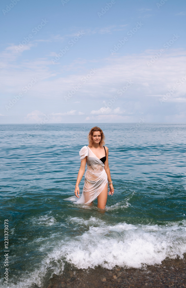 A young beautiful girl in a wet white tunic and with a smartwatch on her wrist comes out of the sea. A foam wave. Azure water. The modern concept of the goddess. Full length A young beautiful girl in a wet white tunic and with a smartwatch on her wrist comes out of the sea. A foam wave. Azure water. The modern concept of the goddess. Full length