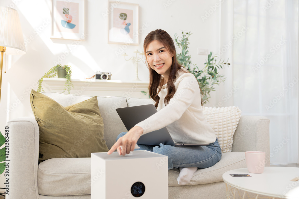 Asian woman adjusts a smart air purifier relaxing at home. ensuring clean and protection from PM2.5 dust, quality of daily life well-being, pollution prevention by innovative technology health care Asian woman adjusts a smart air purifier relaxing at home. ensuring clean and protection from PM2.5 dust, quality of daily life well-being, pollution prevention by innovative technology health care