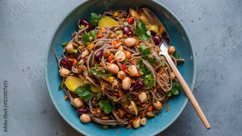 Vibrant vegan soba noodle salad with chickpeas, peanuts, and herbs in a teal bowl, garnished with colorful beans and garnishes on a gray surface