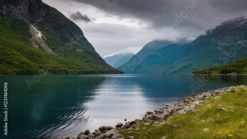 Serene fjord landscape on a rainy overcast summer day, featuring lush green mountains, calm turquoise water, and dramatic cloudy sky reflections.