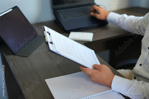 An asian businessman holding pen to write or sign business document and contract sheet while sitting at desk with coffee cup, laptop and working alone in home office, business and office concept.