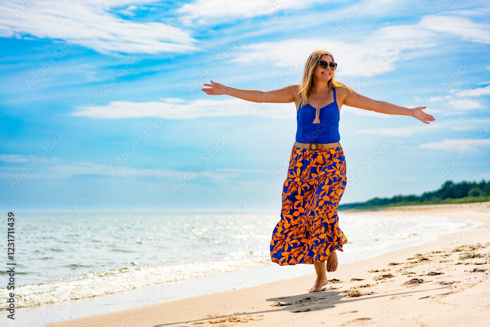 Summertime by sea. Happy middle-aged woman walking with arms stretched out on sandy beach on sunny summer day. Happiness and satisfaction. Front view Summertime by sea. Happy middle-aged woman walking with arms stretched out on sandy beach on sunny summer day. Happiness and satisfaction. Front view