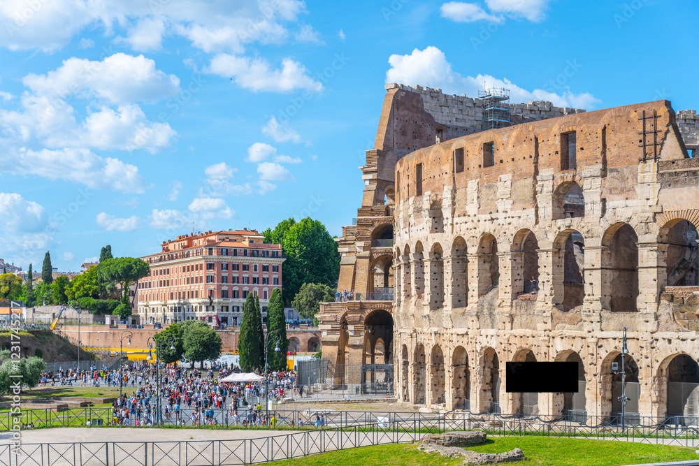 Fototapeta premium Crowds gather near the Colosseum in Rome, Italy, under a bright blue sky. This iconic landmark showcases ancient architecture and attracts tourists year-round with its historical significance.