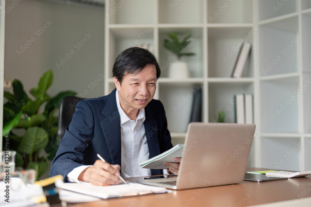 Elderly Asian businessman reviewing documents while taking notes on a laptop in a modern office. Elderly Asian businessman reviewing documents while taking notes on a laptop in a modern office.