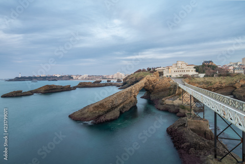Vue depuis le Rocher de la Vierge à Biarritz, capturant l’océan Atlantique et le paysage urbain. Pont emblématique, rochers sculptés par les vagues et horizon côtier offrent une scène pittoresque.