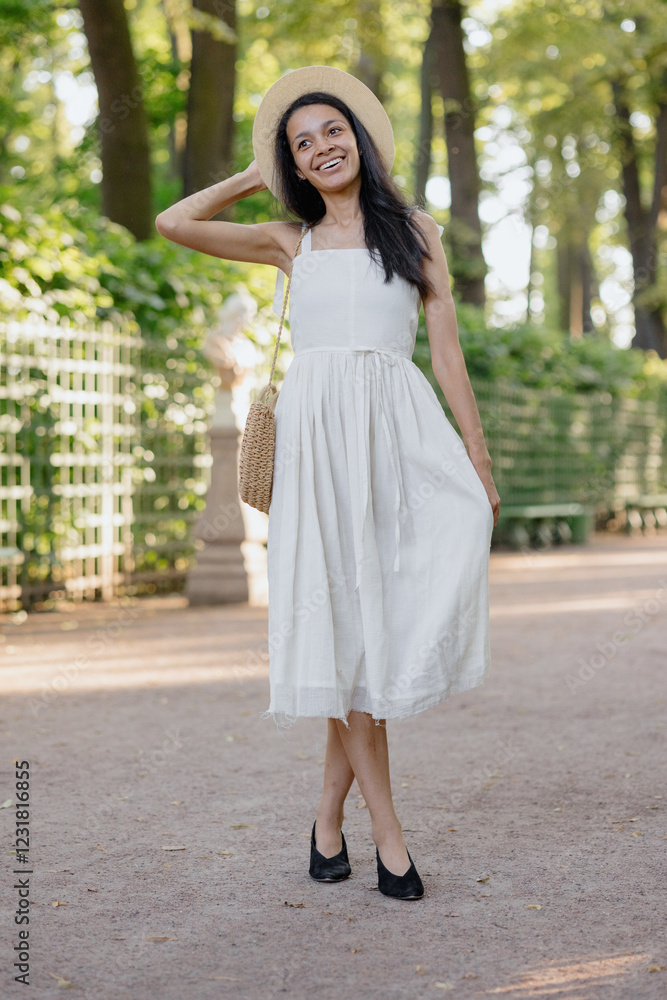 carefree African American woman in white dress and straw hat walks in park and poses against background of green hedge. daytime for relaxation. beautiful female fashion model, sunlight summer time carefree African American woman in white dress and straw hat walks in park and poses against background of green hedge. daytime for relaxation. beautiful female fashion model, sunlight summer time