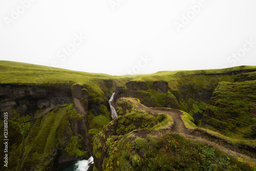 Vue spectaculaire du canyon de Fjaðrárgljúfur en Islande, recouvert d’une mousse verte luxuriante. Une cascade serpente entre les falaises, créant un paysage féerique sous une brume mystique.