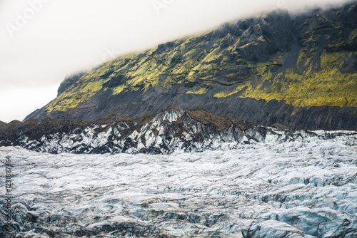 Paysage spectaculaire d’un glacier islandais bordé de montagnes recouvertes de mousse verte. Contraste frappant entre la glace, la roche volcanique et la brume, capturant la beauté brute de l’Islande.