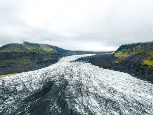Photographie aérienne spectaculaire capturée par drone, mettant en valeur un glacier islandais avec ses stries de glace, ses dépôts de cendres volcaniques et son relief montagneux abrupt. La vue plong