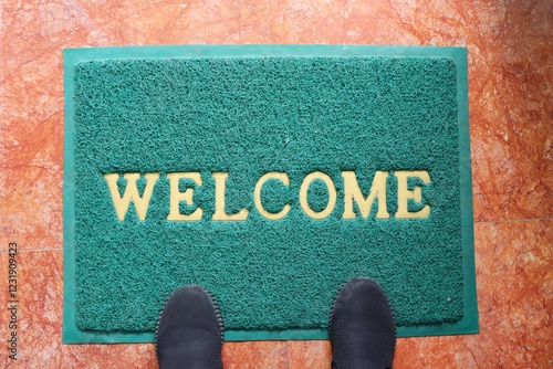 A green welcome doormat with feet wearing black shoes. Interior and object concept.