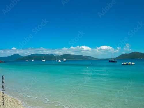 Turquoise waters of the Great Barrier Reef near Green Island with anchored sailboats and yachts against mountainous backdrop. Crystal-clear tropical waters meet coral beach shoreline, Queensland sky
