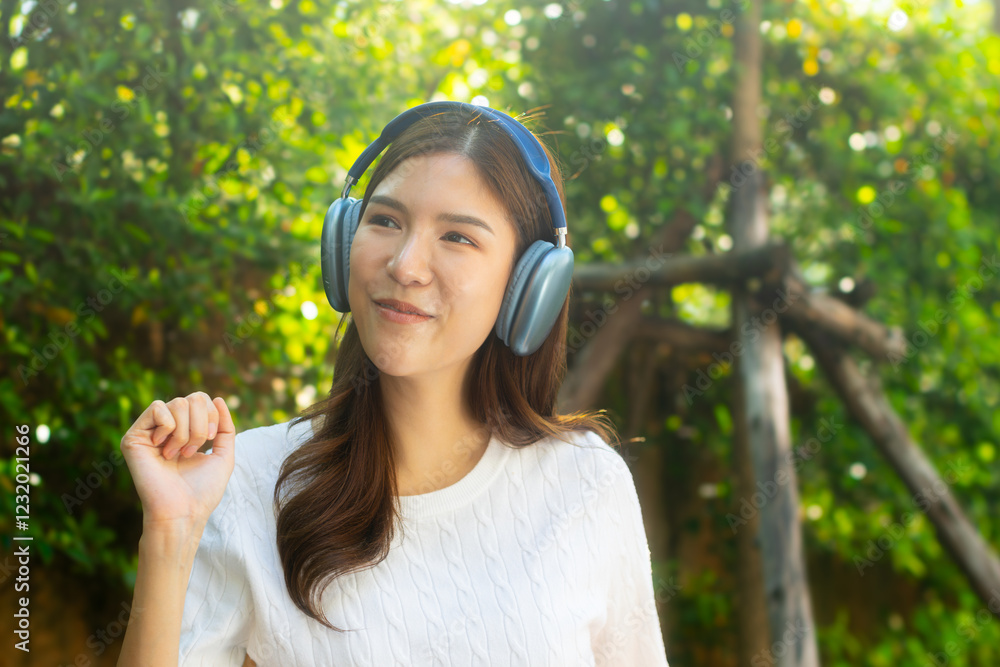 Attractive young Asian woman sits in a park, wearing wireless headphones and smiling, listening to music while relaxing in a tranquil park. Joyful Asian woman feeling relaxed, happy, and stress-free. Attractive young Asian woman sits in a park, wearing wireless headphones and smiling, listening to music while relaxing in a tranquil park. Joyful Asian woman feeling relaxed, happy, and stress-free.