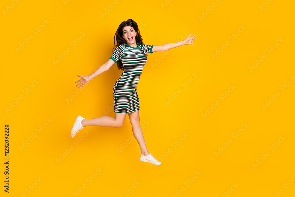 Cheerful young woman jumping joyfully on bright yellow background wearing striped casual dress Cheerful young woman jumping joyfully on bright yellow background wearing striped casual dress