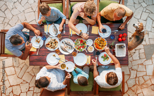 Top view of cheerful family gathered around outdoor dining table enjoying Mediterranean meal with fresh fish, vegetables, and salads, vibrant dishes on rustic terrace, summer vibe, togetherness.