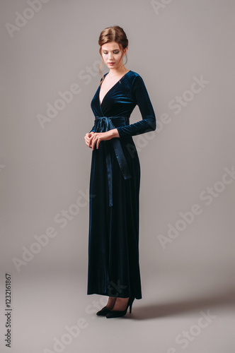 Wrap maxi velour evening dress in navy blue. Long sleeve velvet gown with bow knot. Studio portrait of elegant young woman on high heels, fashionable outfit for an event. Romantic brunette lady