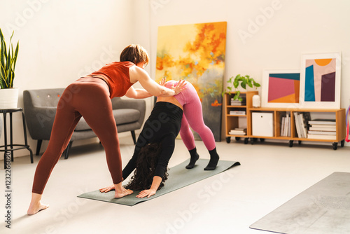 A woman in a red athletic outfit helps another woman in black and pink adjust her posture in a downward-facing dog yoga pose on a mat in a peaceful studio with abstract artwork and plants photo