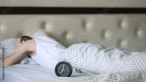 Teenager kid boy lies on bed waking up in morning and gets angry at alarm clock covers his ears with a pillow