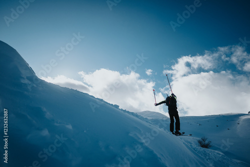 A skier ascends a snow-covered slope under a bright blue sky. The scene captures the adventurous and invigorating spirit of outdoor winter mountain sports in pristine natural surroundings. photo