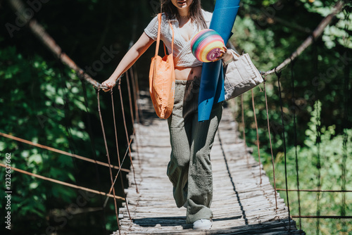 A woman confidently crosses a rustic wooden bridge holding a colorful yoga mat and bag, enjoying a peaceful day in nature. photo