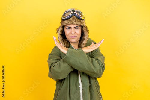 Young beautiful aviator woman wearing vintage pilot helmet with glasses and jacket over yellow background Rejection expression crossing arms doing negative sign, angry face photo