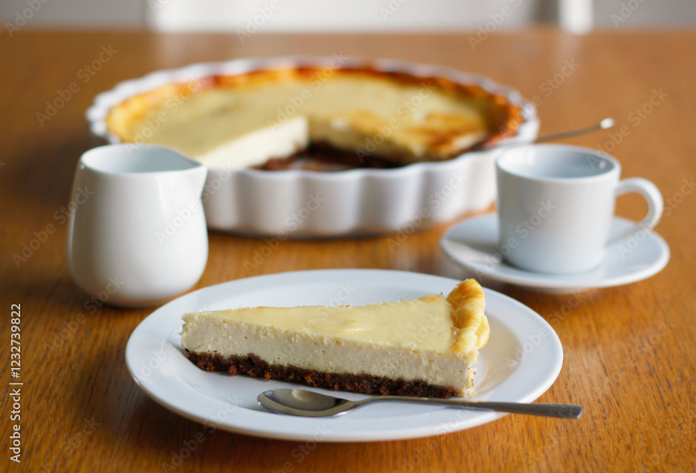 Piece of homemade cheesecake on a white plate on wooden table, a cup of black coffee with saucer, a cup of milk and a baking dish in the background. Coffee nad cake, side view, selective focus. Piece of homemade cheesecake on a white plate on wooden table, a cup of black coffee with saucer, a cup of milk and a baking dish in the background. Coffee nad cake, side view, selective focus.