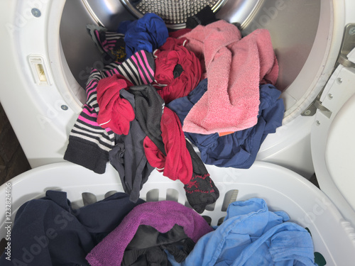 Photos Closeup of a variety of washed and dried clothes spilling out of a front loading tumble dryer drum, straight into a laundry basket on the floor