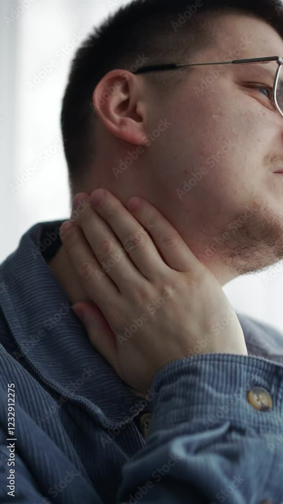 Young man experiencing neck pain while holding his throat in a thoughtful expression indoors Young man experiencing neck pain while holding his throat in a thoughtful expression indoors