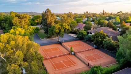 Aerial view of red clay tennis courts in a quiet Australian neighborhood, bordered by eucalyptus trees and modern suburban houses, softly illuminated by the setting sun