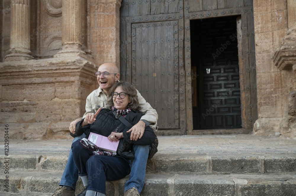 Senior couple laughing and embracing while sitting on the stone steps of a historic church during a cultural trip, enjoying their travel experience in Spain Senior couple laughing and embracing while sitting on the stone steps of a historic church during a cultural trip, enjoying their travel experience in Spain