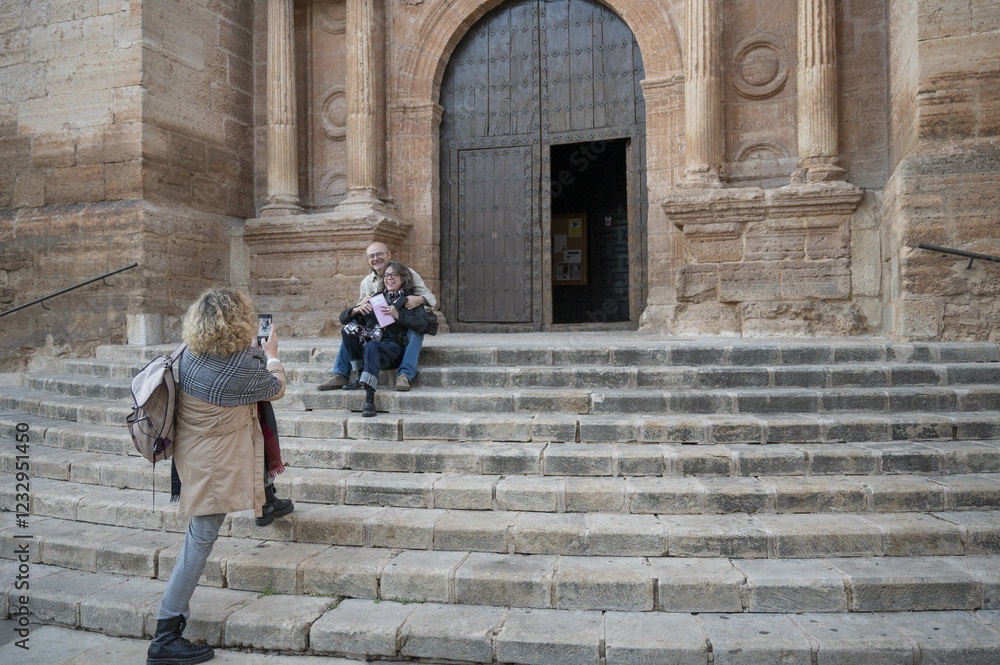 Senior tourists capturing memories in front of a historic church, as a woman takes a picture of her two friends sitting on the stone steps, enjoying a cultural trip in Spain