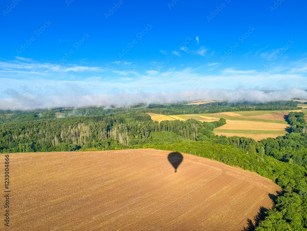 Obraz premium The shadow of the hot air balloon falls on the fields of wheat, at the level of the clouds. Europe, Czech Republic